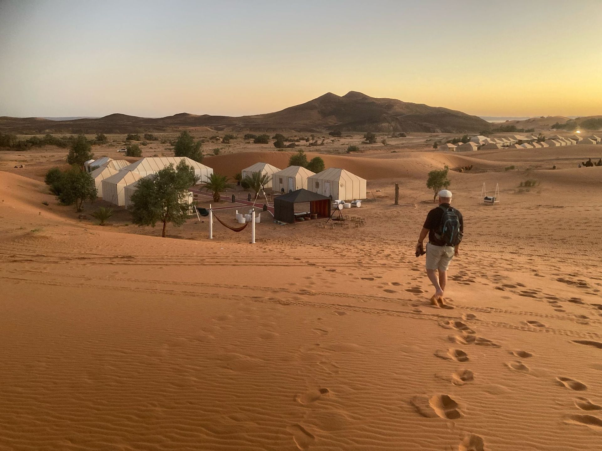 A man is walking through a desert with tents in the background.