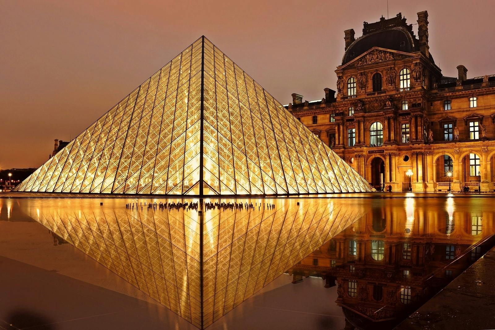 Louvre Pyramid at night, reflecting in water; golden light illuminates the glass structure and nearby building.