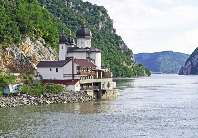 White building with domes on a rocky cliffside, overlooking a wide river. Green trees and hills in background.