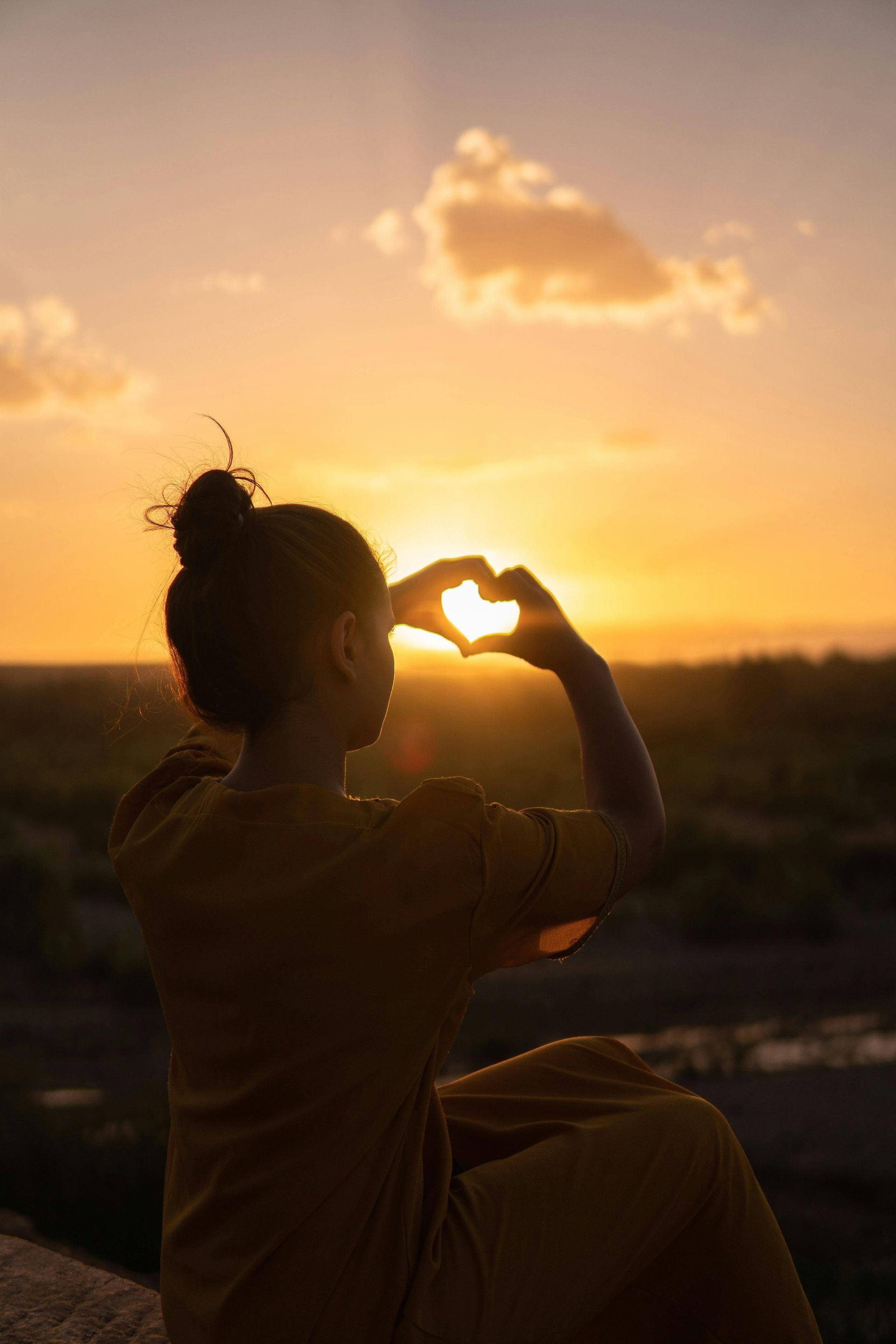 A woman is making a heart shape with her hands at sunset.