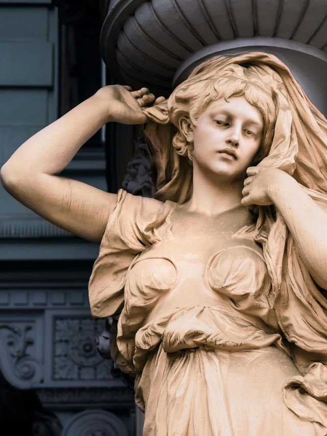 Statue of a woman with long hair, holding her hair up, with a classical building in the background.
