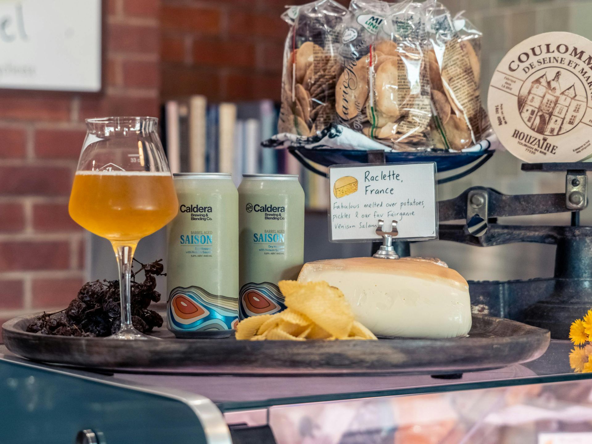 A tray with beer, chips, cheese, and cookies sits on a counter, with scales and a brick wall background.