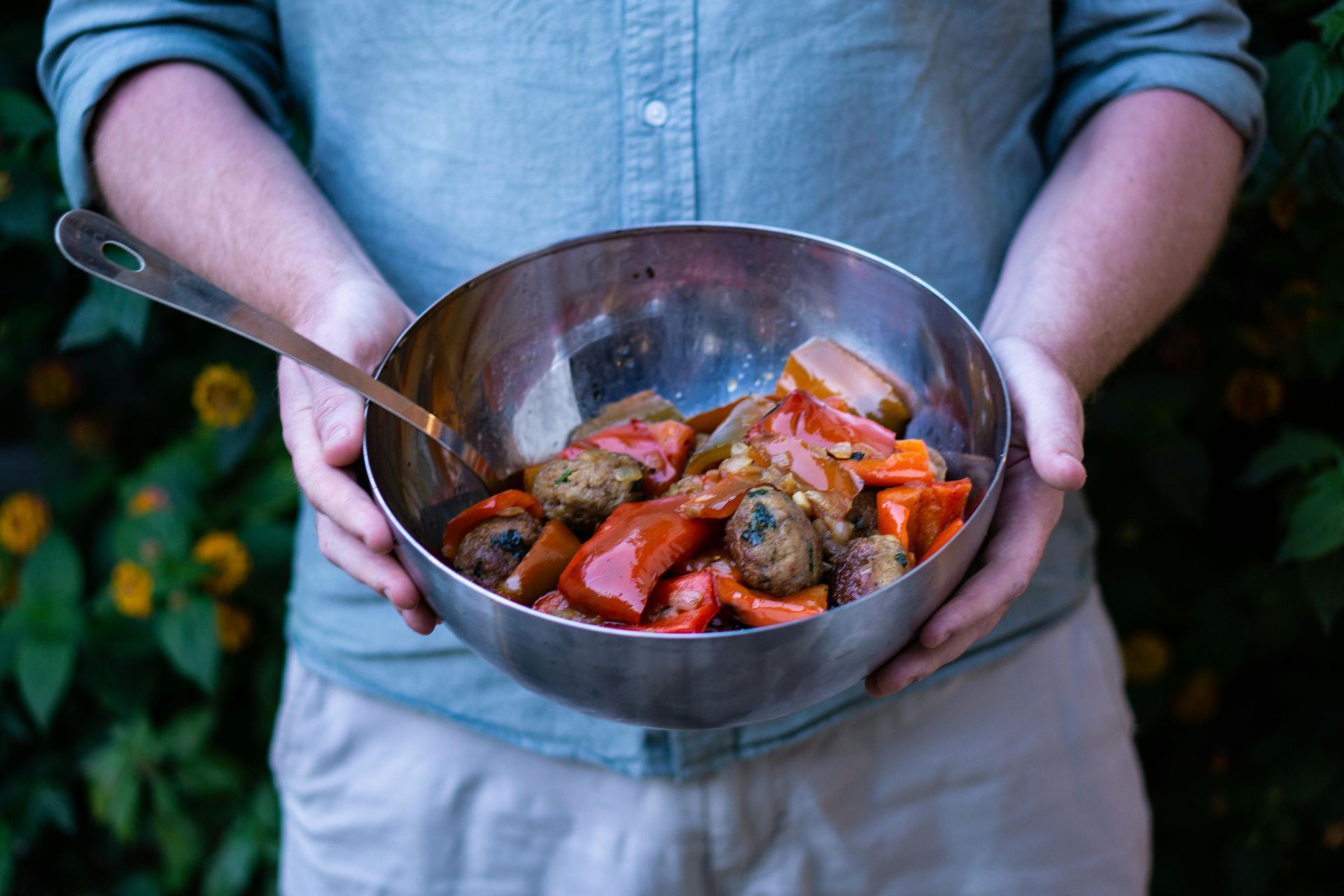 Person holding a large metal bowl of stew, spoon inside. Includes vegetables and meatballs, against a leafy backdrop.