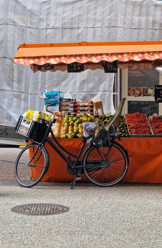 Black bicycle in front of a fruit stand with an orange awning. Produce displayed, cobblestone street.