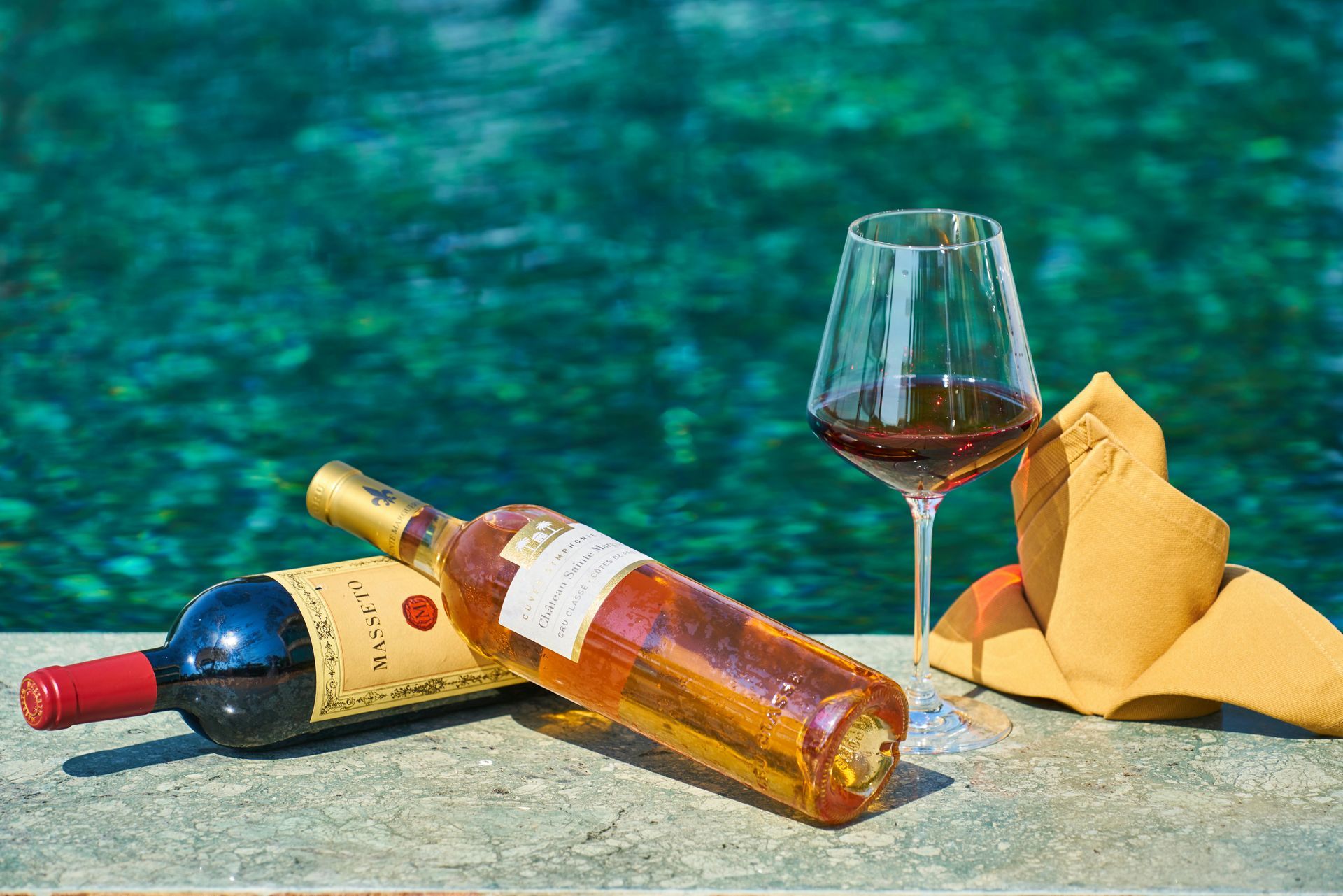 A bottle of wine and a glass of wine are sitting next to a pool.