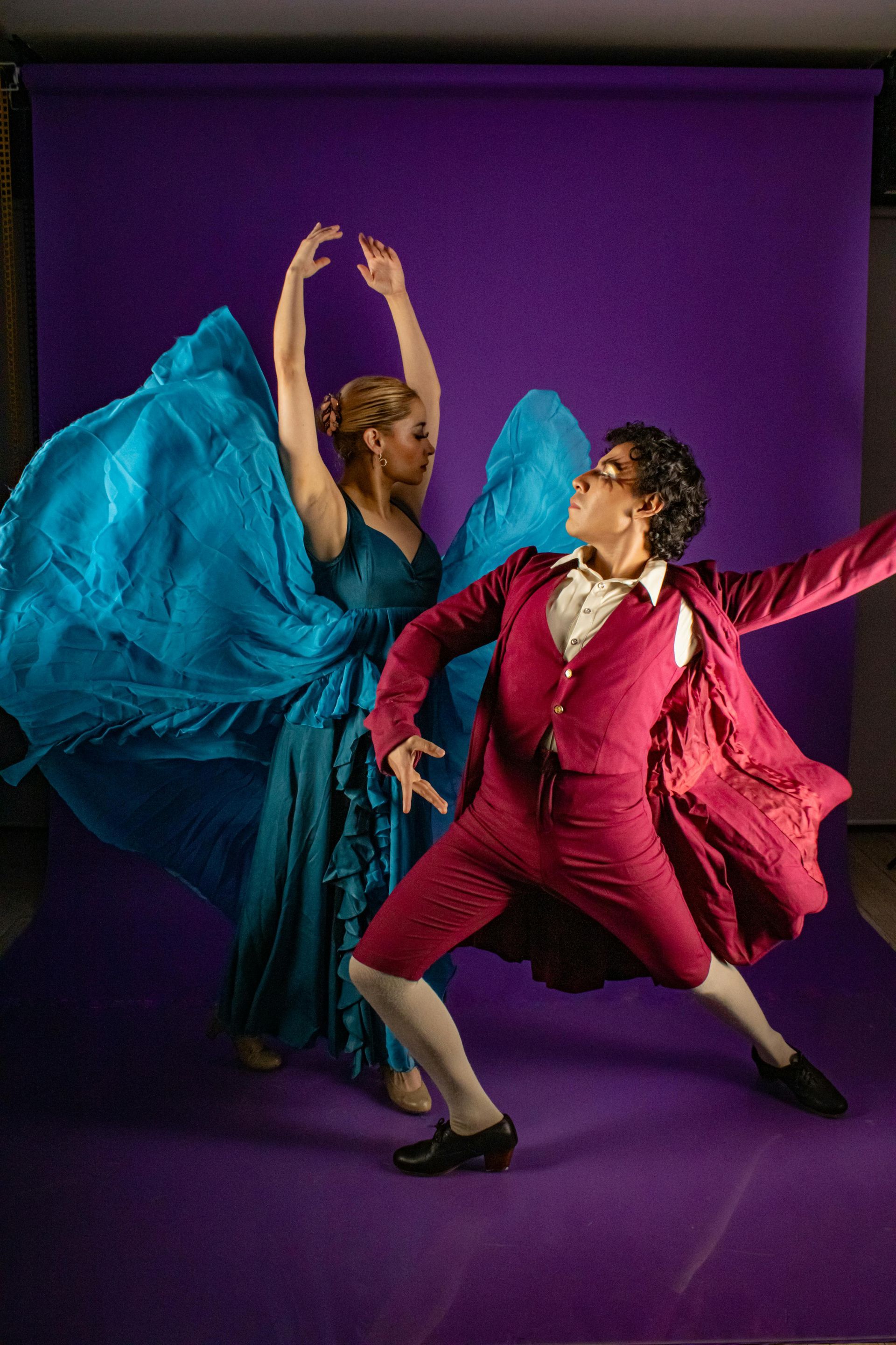 Two dancers on a purple backdrop. One in turquoise dress, arms raised; one in red suit, arm extended.