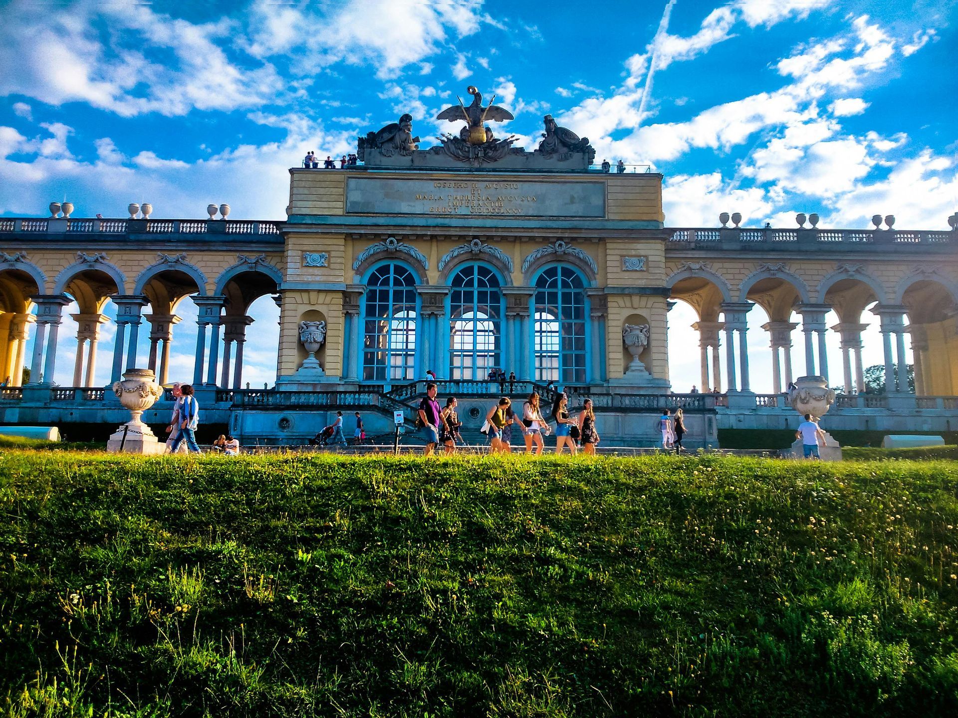 Yellow Schönbrunn Palace structure with arched columns, people, and a blue sky with clouds.
