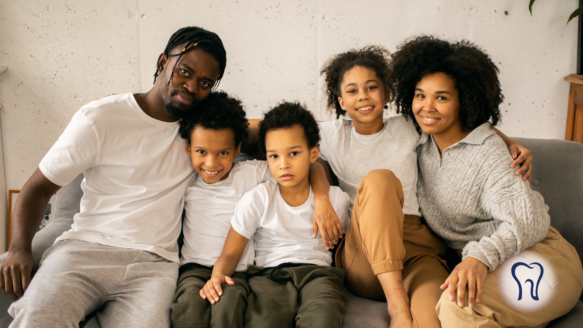 Family of five smiling, posing on a sofa. They wear light-colored clothing and look toward the camera.