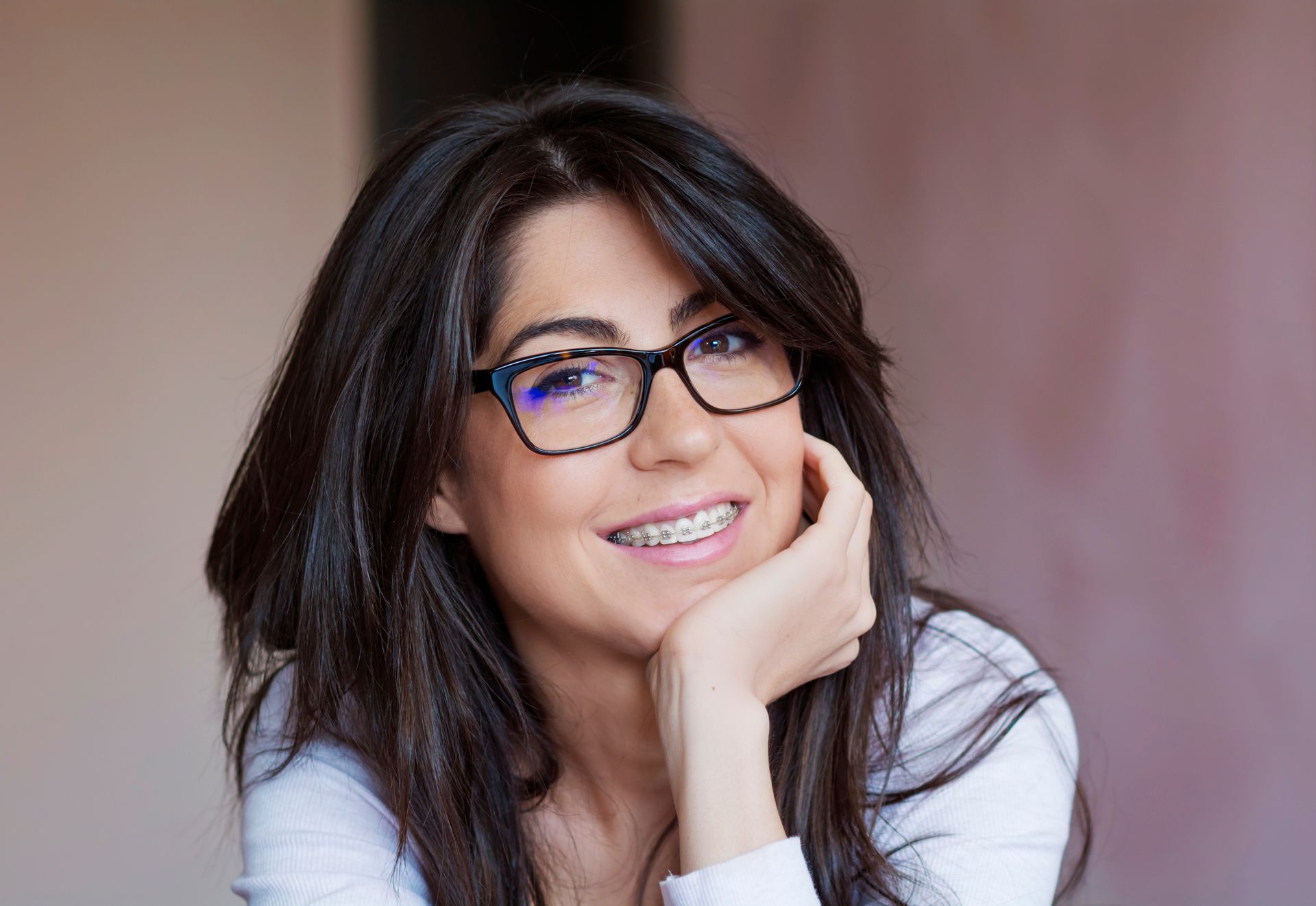 A woman wearing glasses and braces is smiling with her hand on her chin.