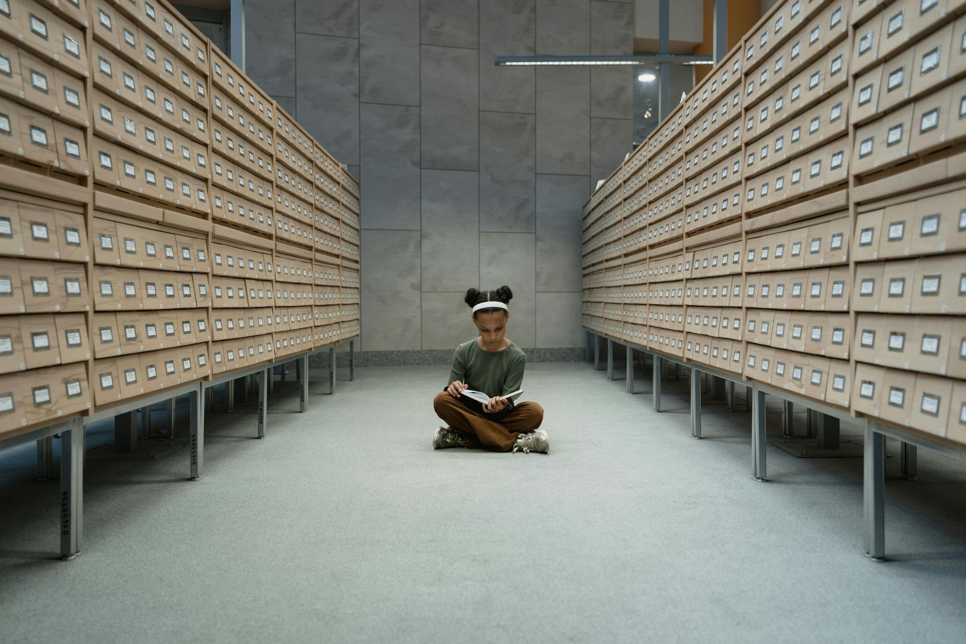 A little girl is sitting on the floor in a library reading a book.