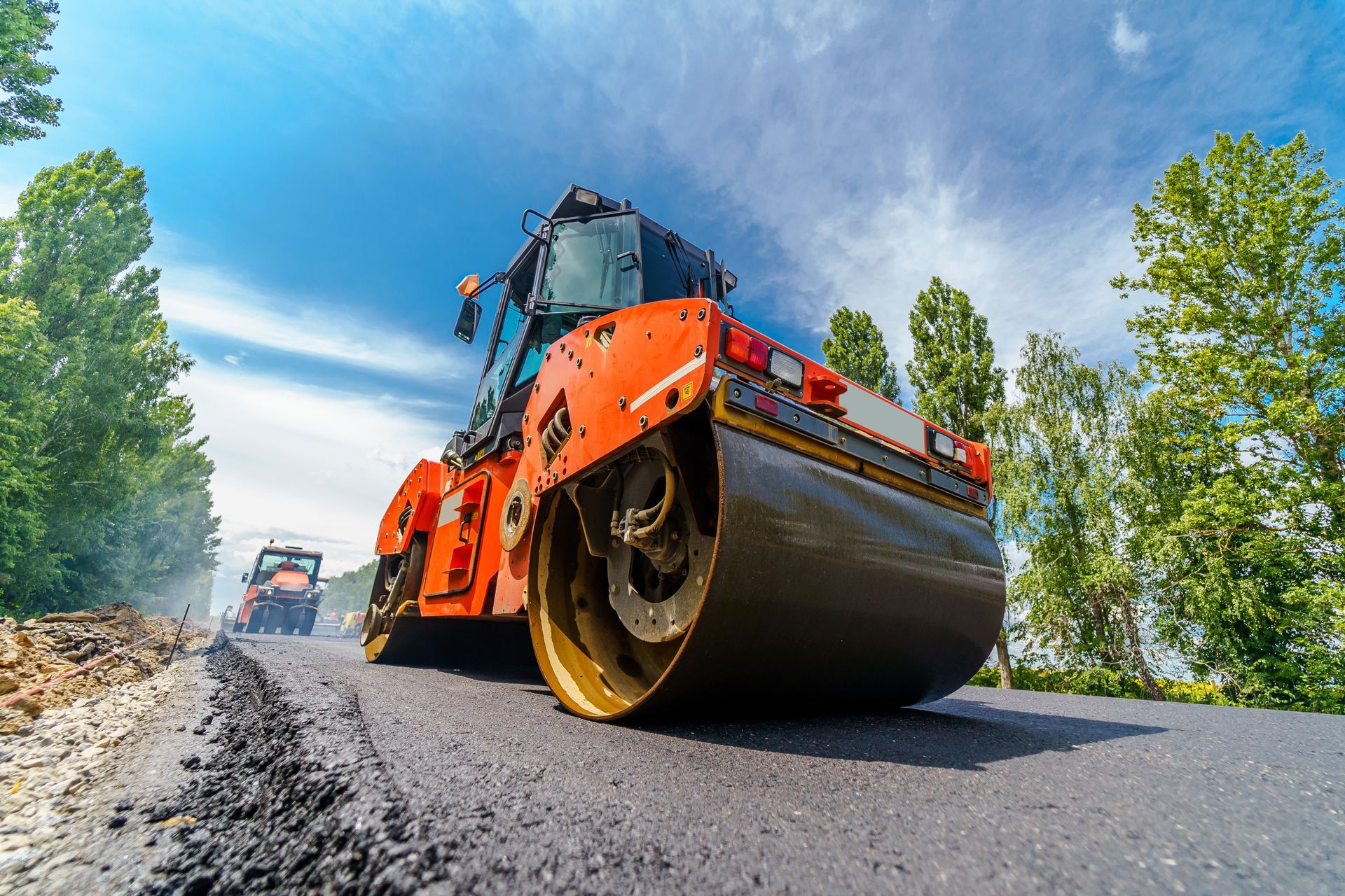Road roller compacting asphalt on a newly paved road under a bright blue sky.