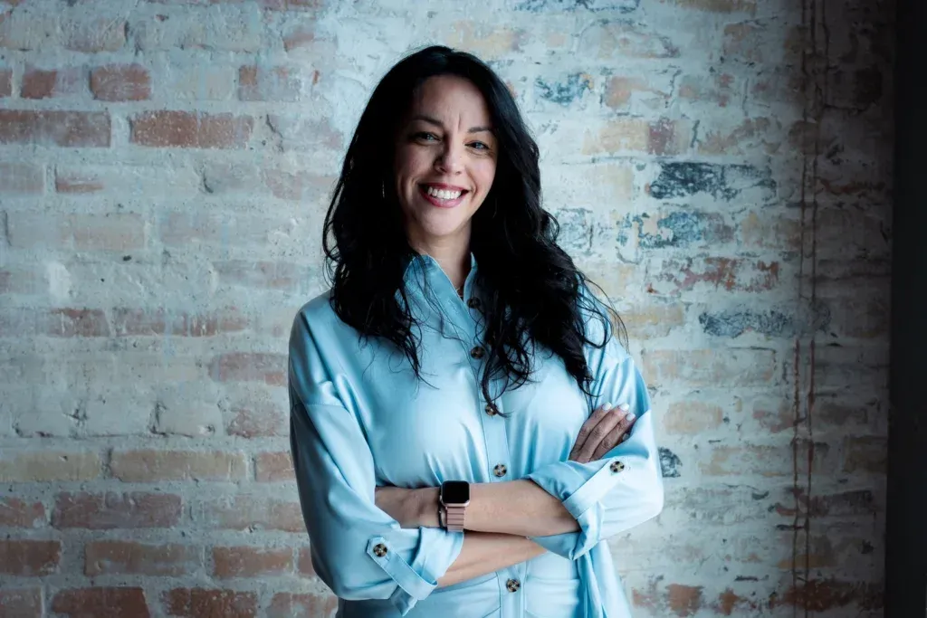 A woman in a blue shirt is standing in front of a brick wall with her arms crossed.