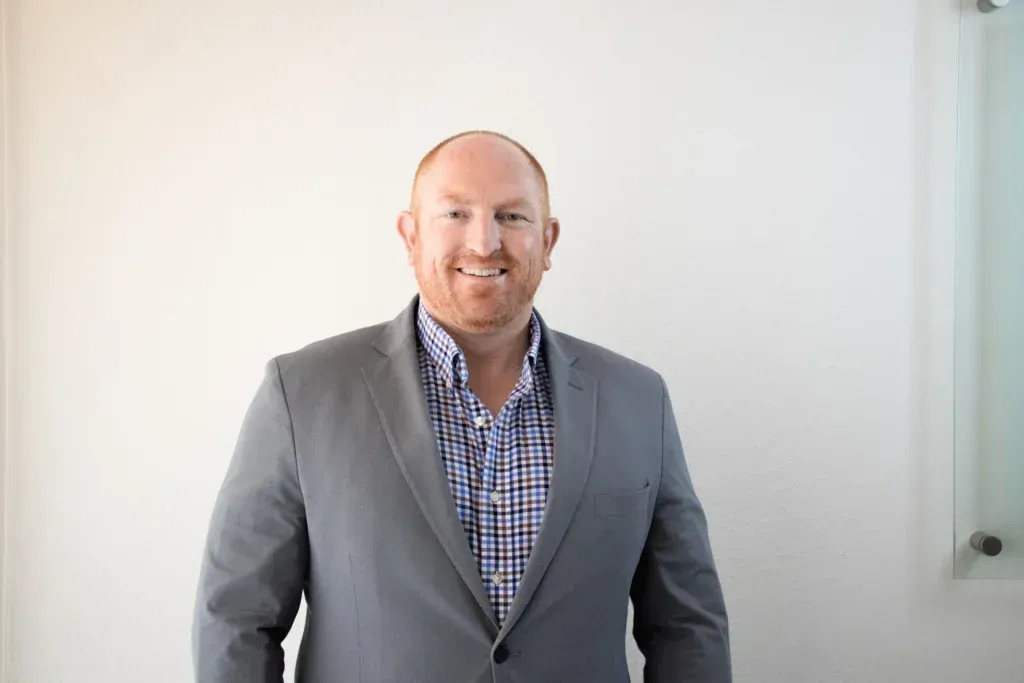 A man in a suit and plaid shirt is standing in front of a white wall.