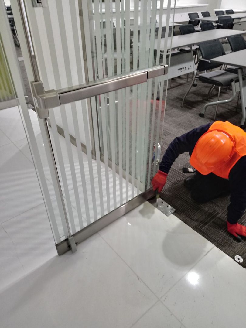 Un trabajador con chaleco de seguridad naranja y guantes se arrodilla para instalar un componente metálico en el marco de una puerta de cristal en una oficina.