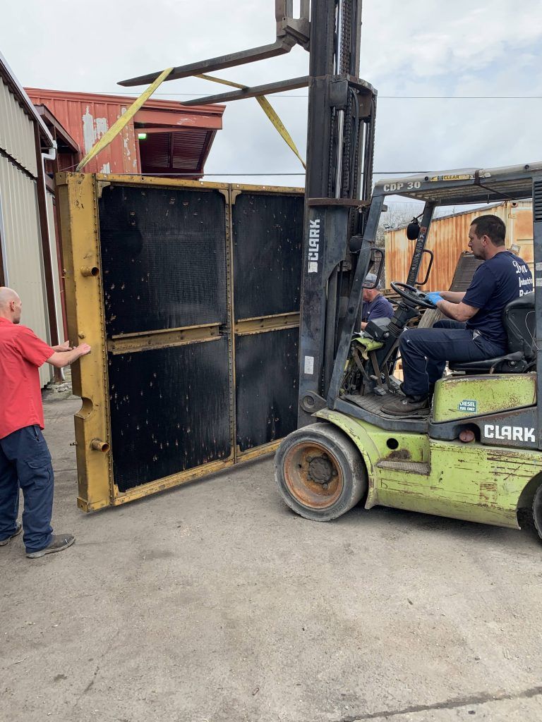 A forklift lifts a large, black rectangular object as two men assist, one guiding it. Outdoor setting.