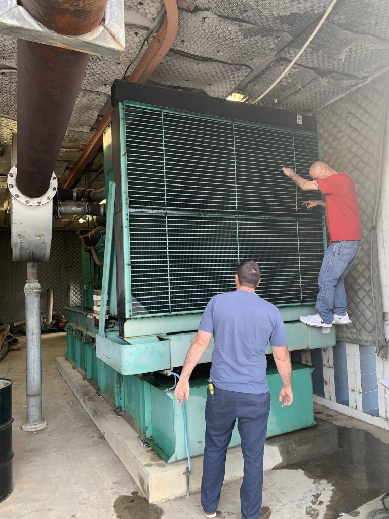 Two men inspecting a large green cooling tower. One man stands at the base, the other is on a ledge, inspecting the tower.