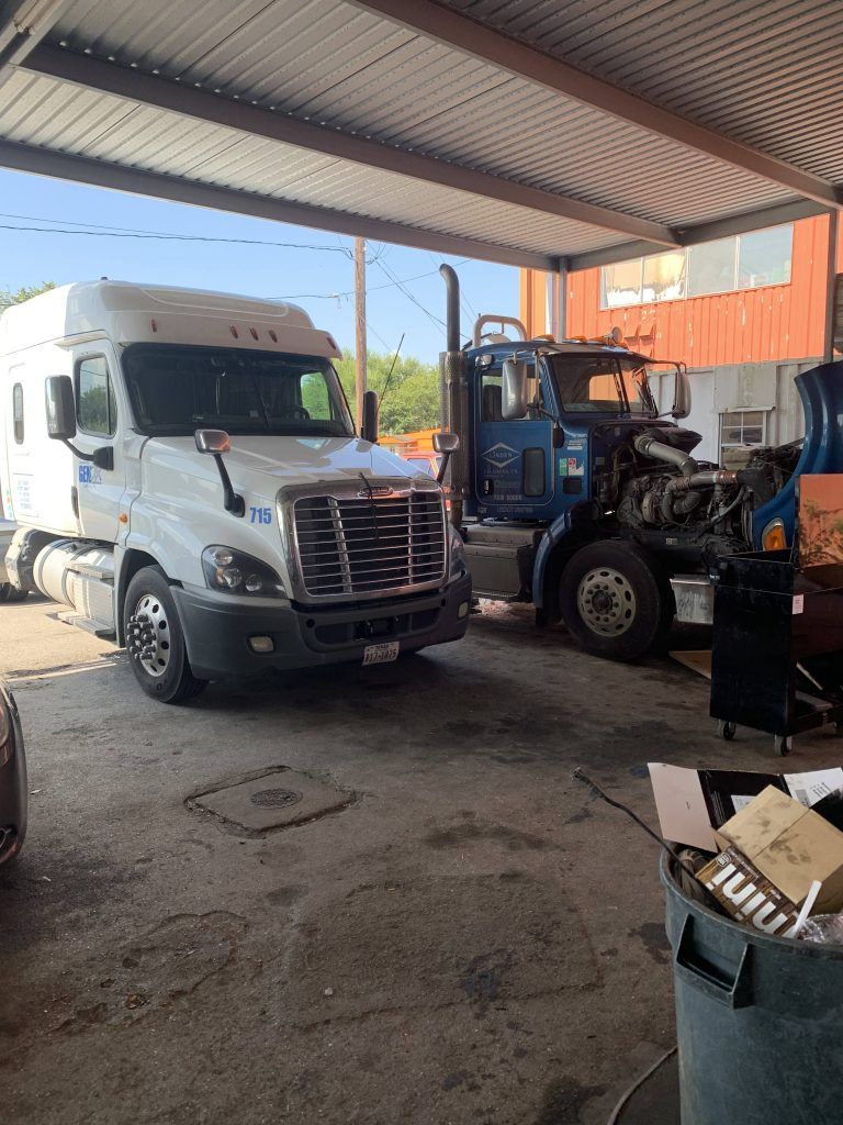 Two semi-trucks under a metal awning, one white and one blue, the latter with open hood.