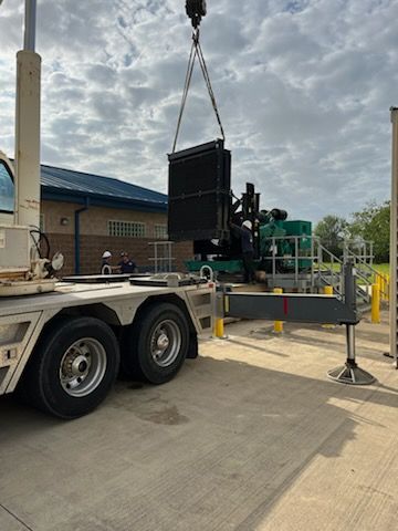 A crane lifting a large black radiator to a generator on a truck bed, outdoors.