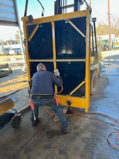 Man cleaning a large black radiator held by a yellow forklift in an outdoor setting.