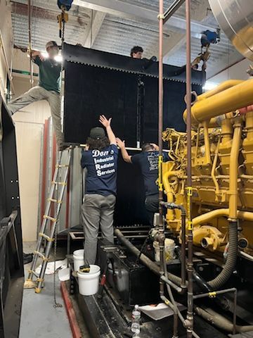 Workers installing a large black industrial heater. Setting is a utility room with yellow pipes, and a ladder.