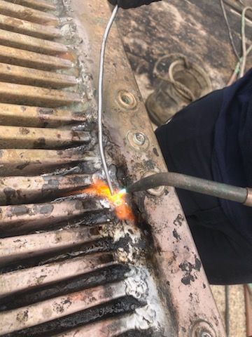A person brazing a metal part with a torch and wire. The part has ridges and holes.