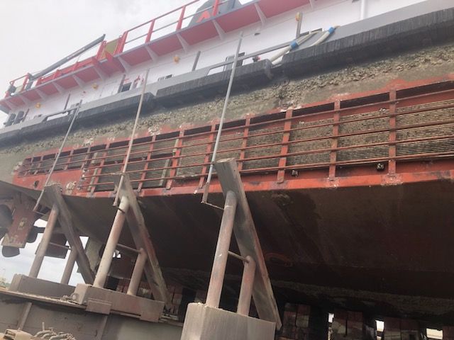 A ship's hull on supports during repair work. Sections of the side are missing, exposing inner structure.