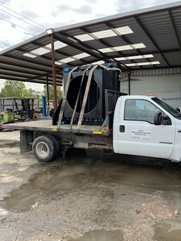 Black industrial equipment secured on a white flatbed truck parked under a metal awning.