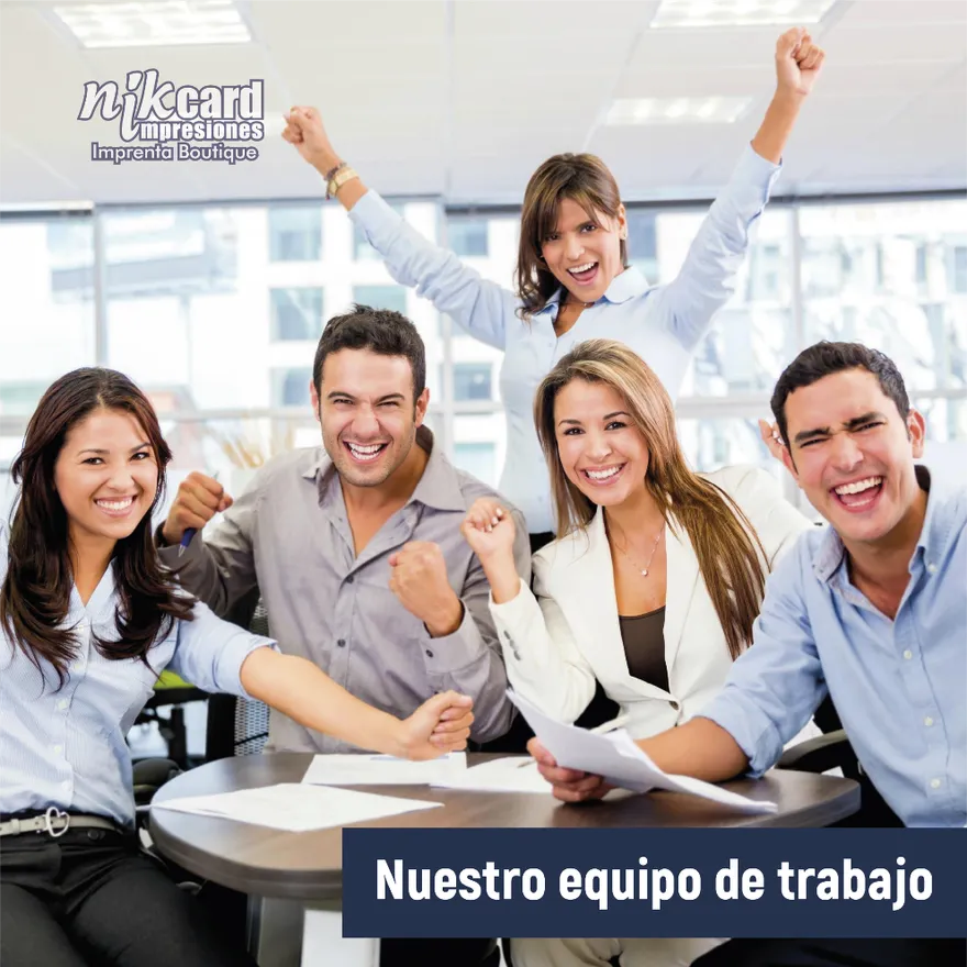 Un equipo de cinco compañeros de trabajo celebra el éxito, sonriendo y levantando los brazos alrededor de una mesa con papeles.