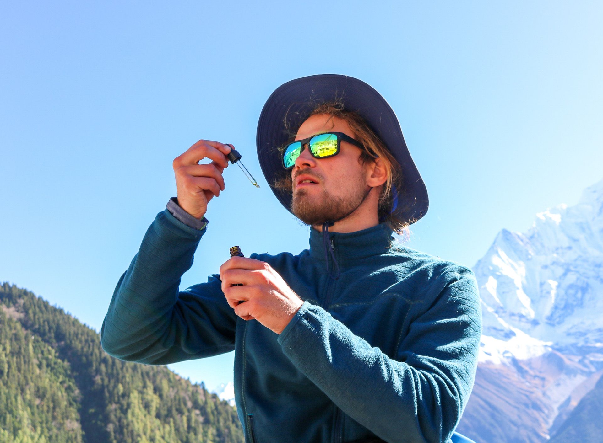 Young man with a beard wearing a fleece, a hat and sunglasses taking CBD oil from a dripper