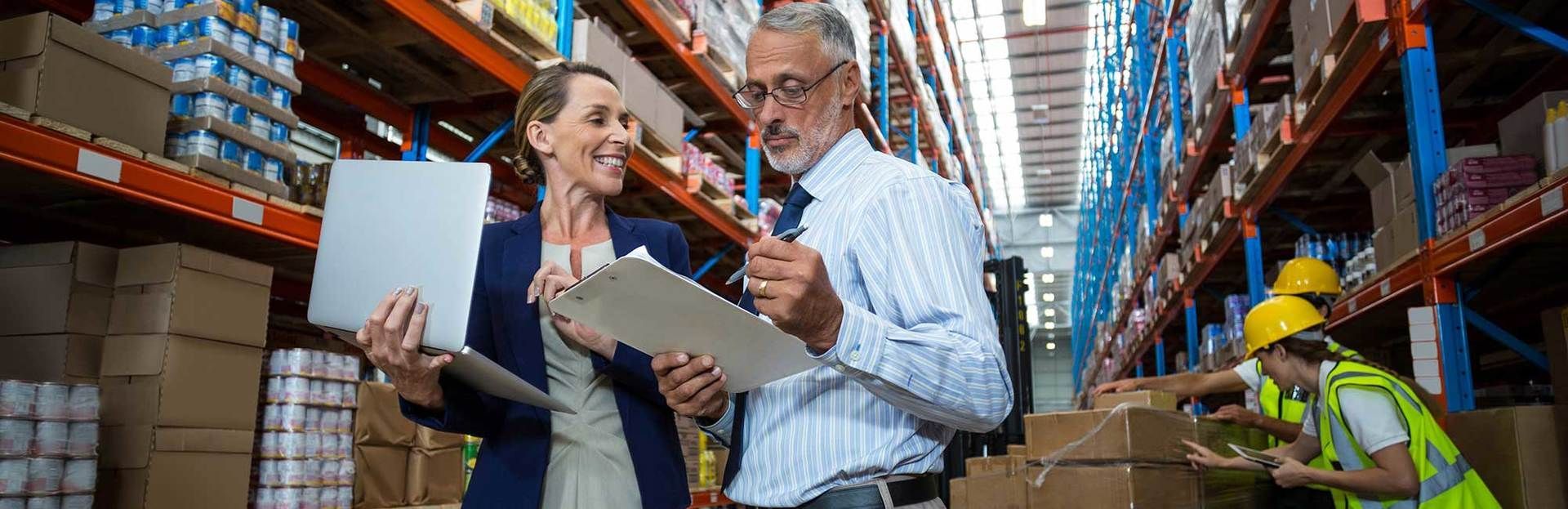 A man and a woman are standing in a warehouse looking at a clipboard.