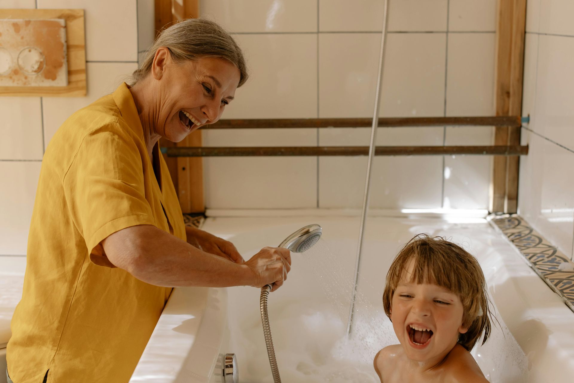 Woman laughs, spraying water on a child in a bathtub. Bathroom setting.