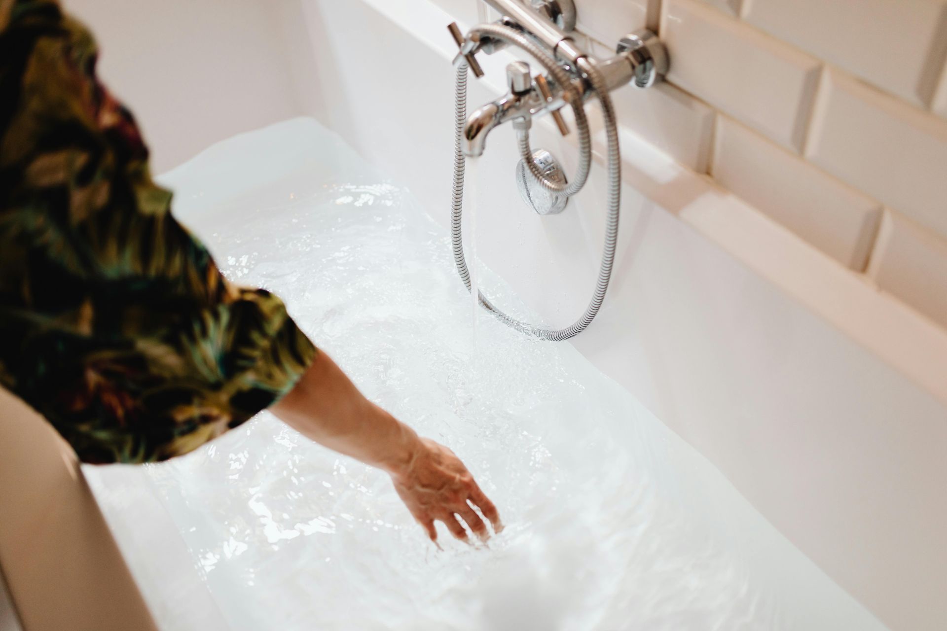 A hand in patterned sleeve reaches toward a bathtub filled with water; bathroom setting with white tiles.