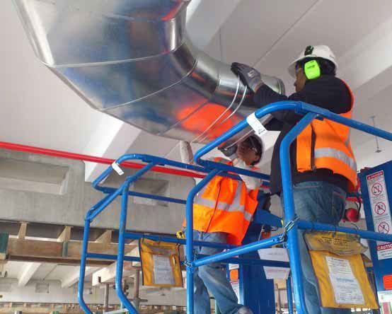 Two workers on a lift are installing a large, duct. 
