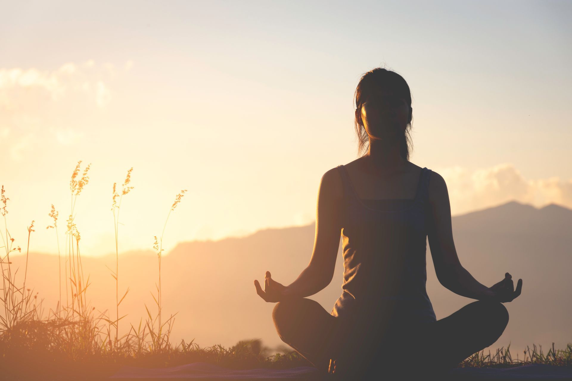 A woman is sitting in a lotus position in front of a mountain at sunset.