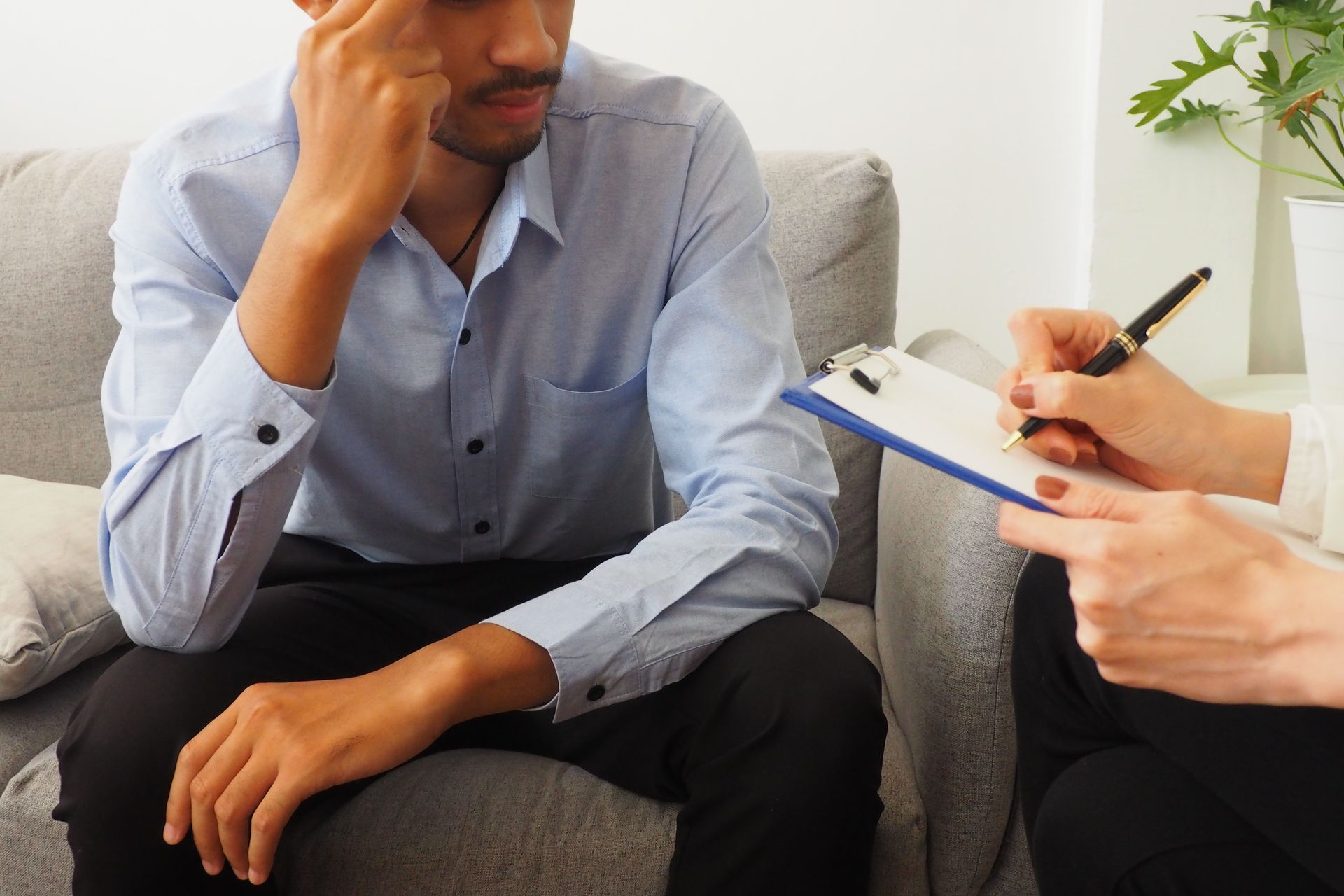 A man is sitting on a couch while a woman writes on a clipboard.