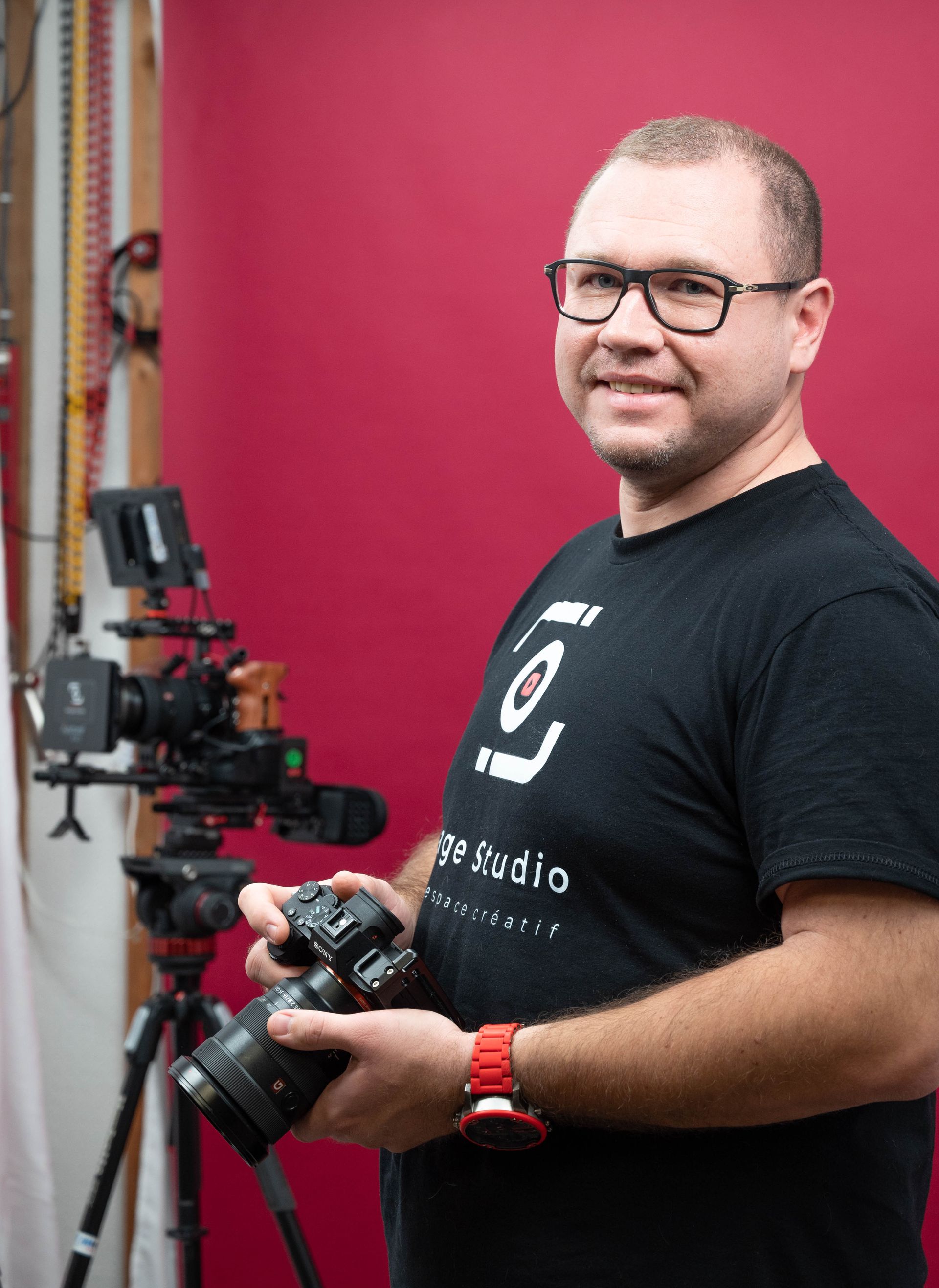 Un homme portant des lunettes tient un appareil photo devant un fond rouge.