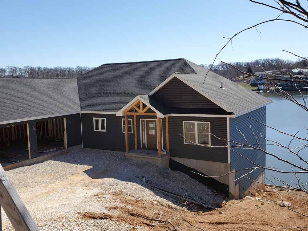 Dark-sided house being built on a waterfront with gray roof, windows, and light gravel landscaping.