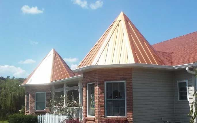 House with red roof, two copper-colored conical roof turrets, brick and beige siding, against a blue sky.