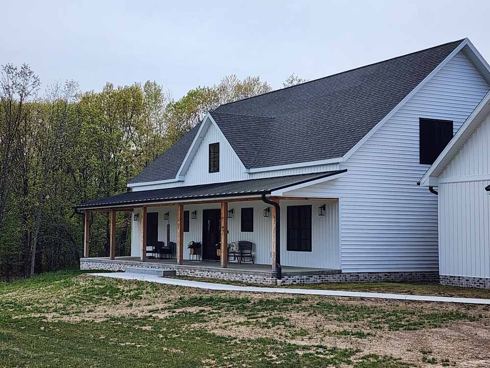 White farmhouse with black roof and porch. Green lawn and trees in background.