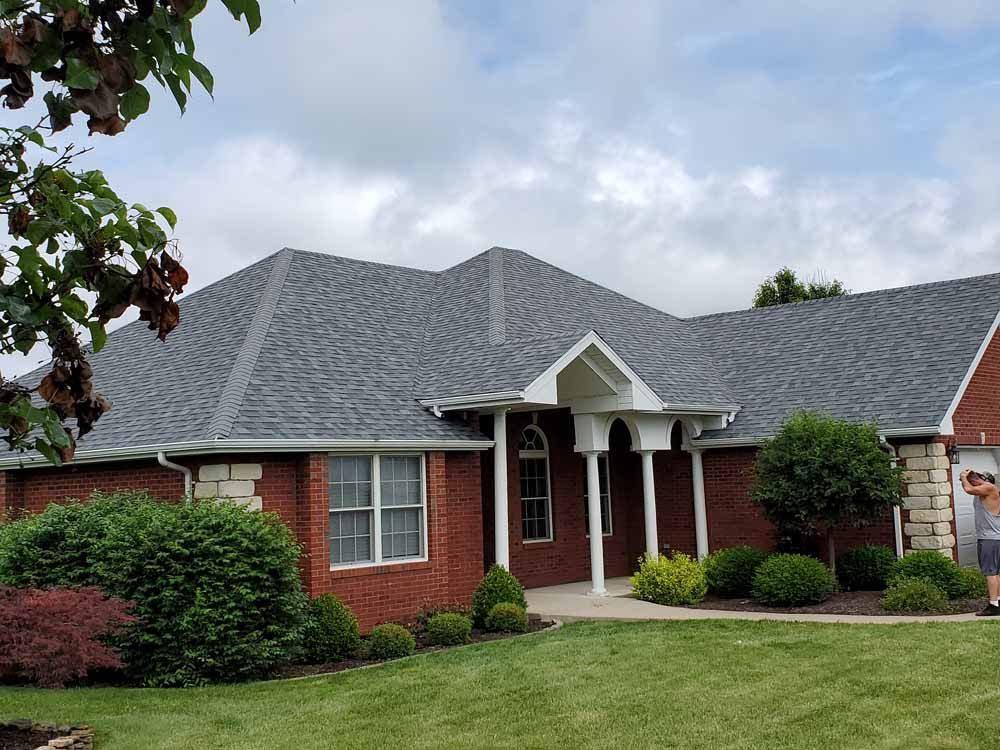 Red brick house with grey shingled roof, green lawn, landscaping, and cloudy sky.