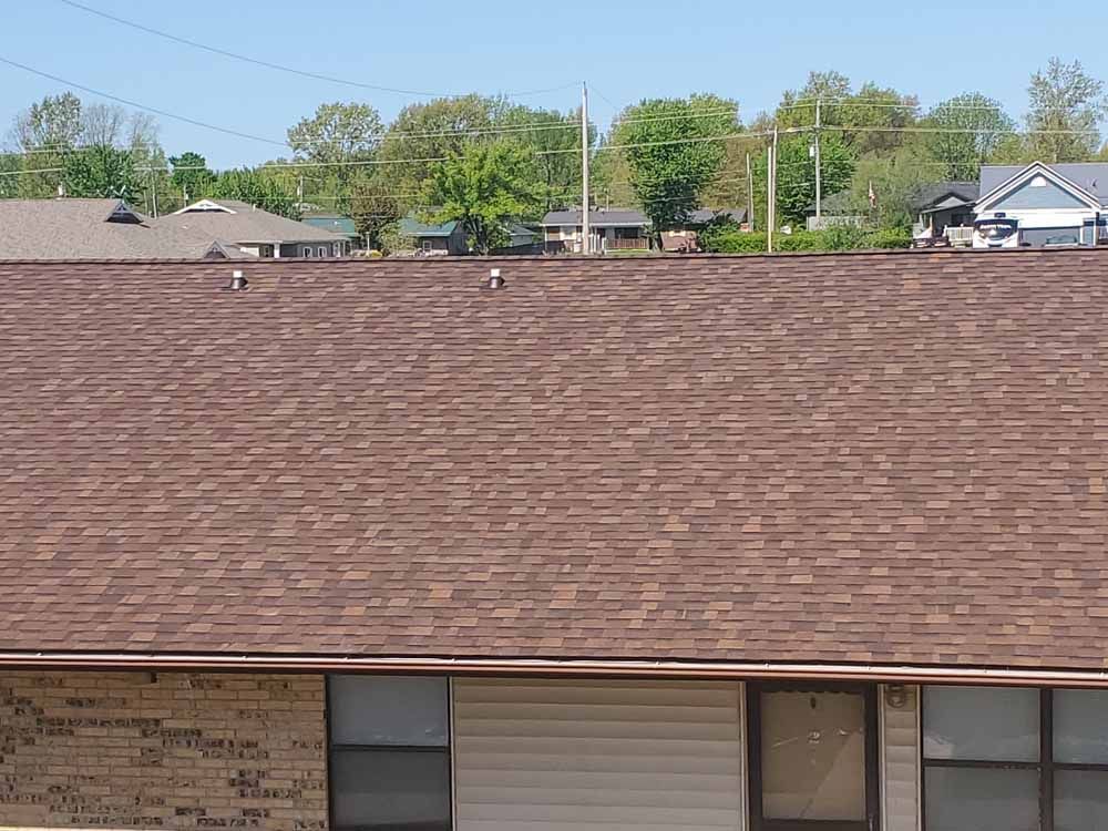 Brown shingled roof with a background of houses, trees, and a blue sky.