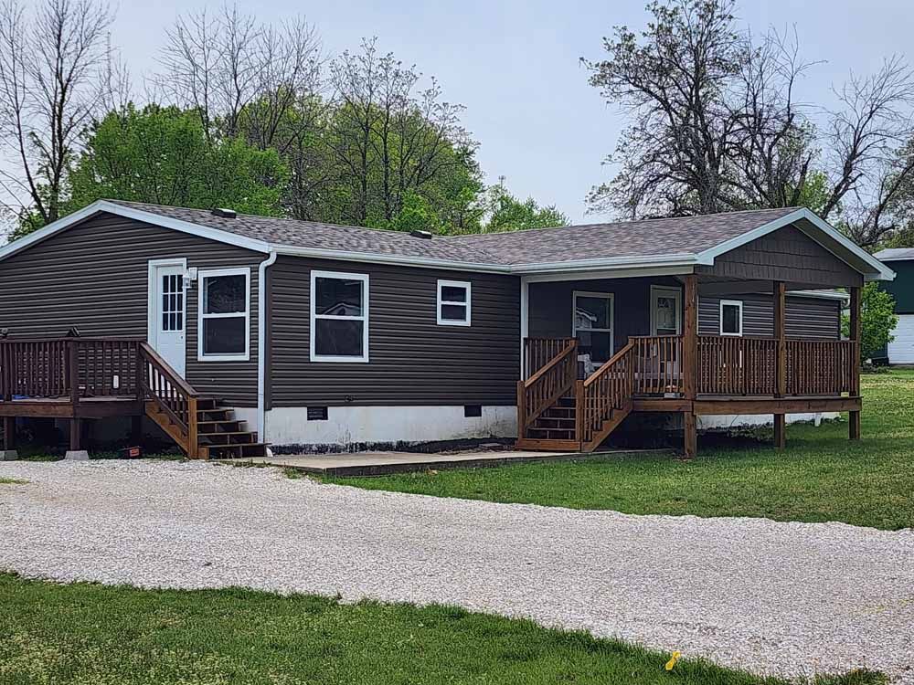 Brown-sided house with wooden porches and steps, gravel driveway, and green grass.