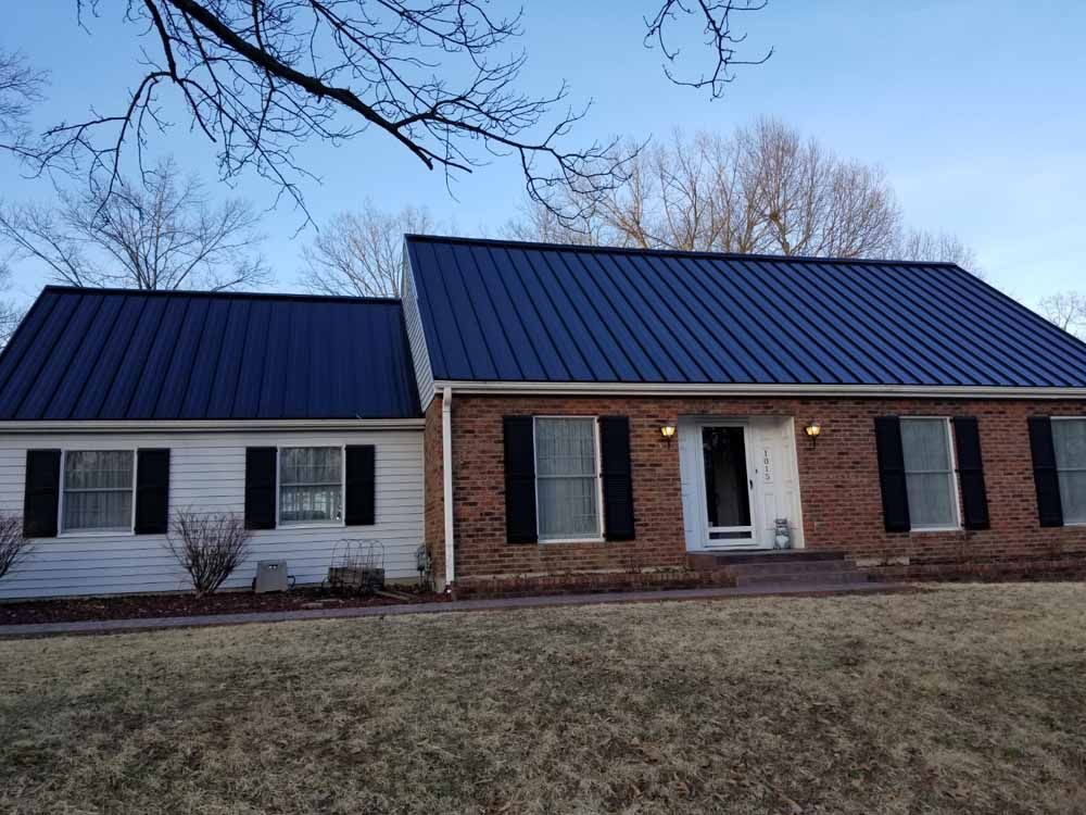 House with dark blue metal roof, brick and white siding, black shutters, and bare trees.