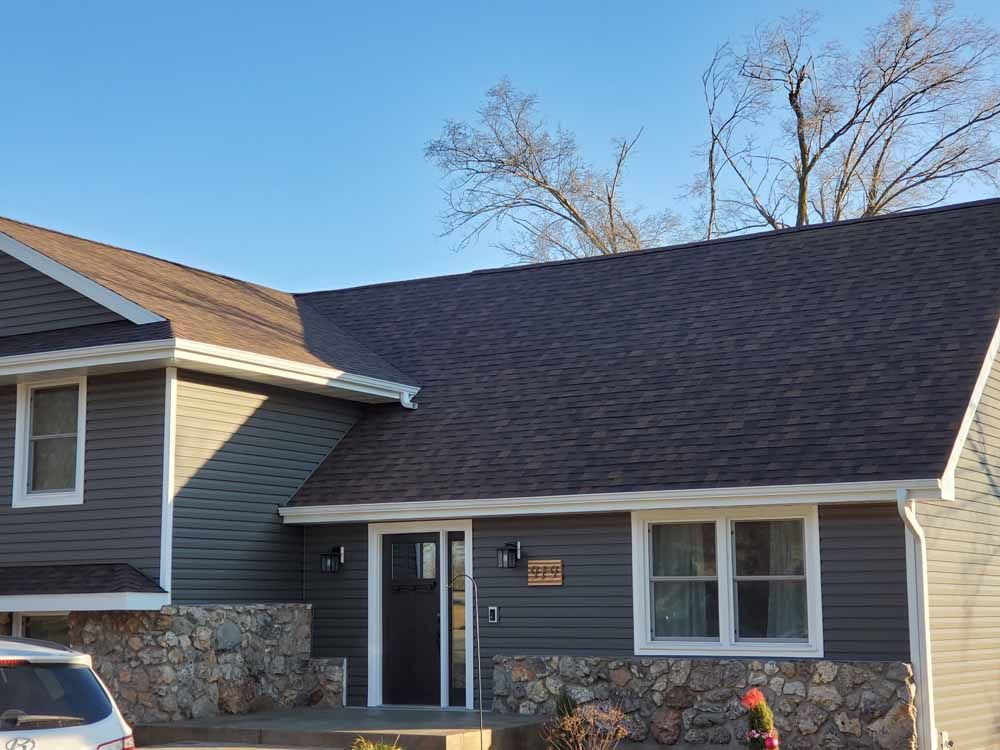 A gray-sided house with a dark roof and a stone foundation, under a clear blue sky.