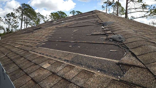 Damaged asphalt shingle roof with missing sections, outdoors with trees in the background.
