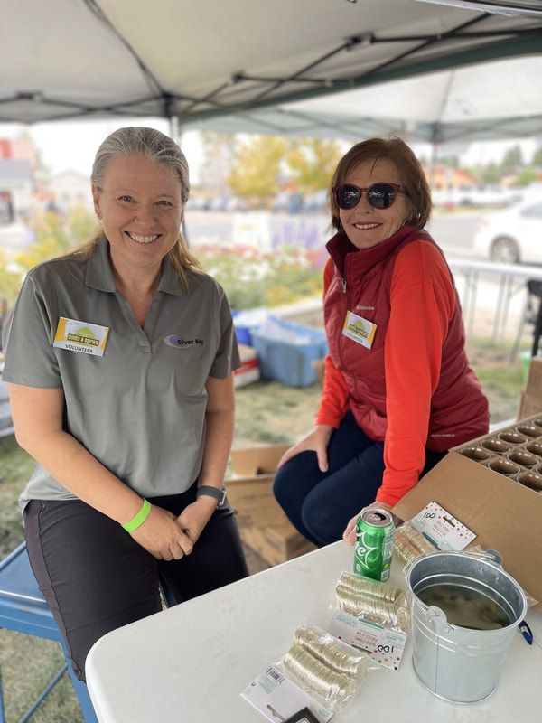 Two women smile at a farmers market booth, offering seed packets. One wears gray, the other red.