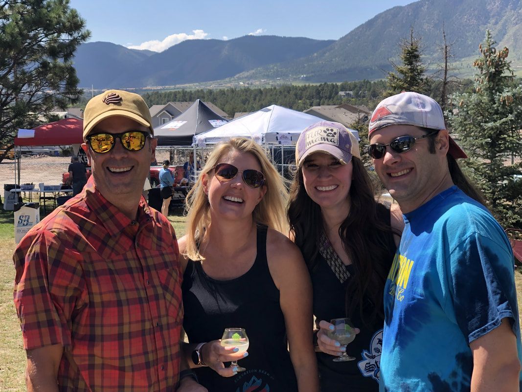 Four smiling people, sunglasses, hats, outdoors in front of mountains and tents.