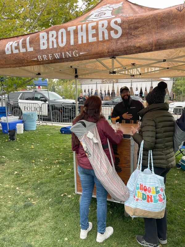 People at a Bell Brothers Brewing booth. Two customers at the counter, one carrying a yoga mat. A man is serving behind the counter.
