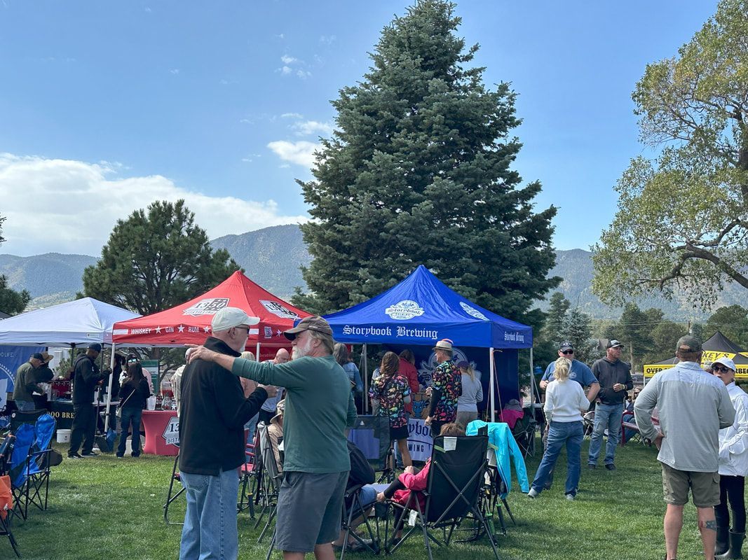 People at an outdoor event near tents and a large evergreen tree; mountains in the background.
