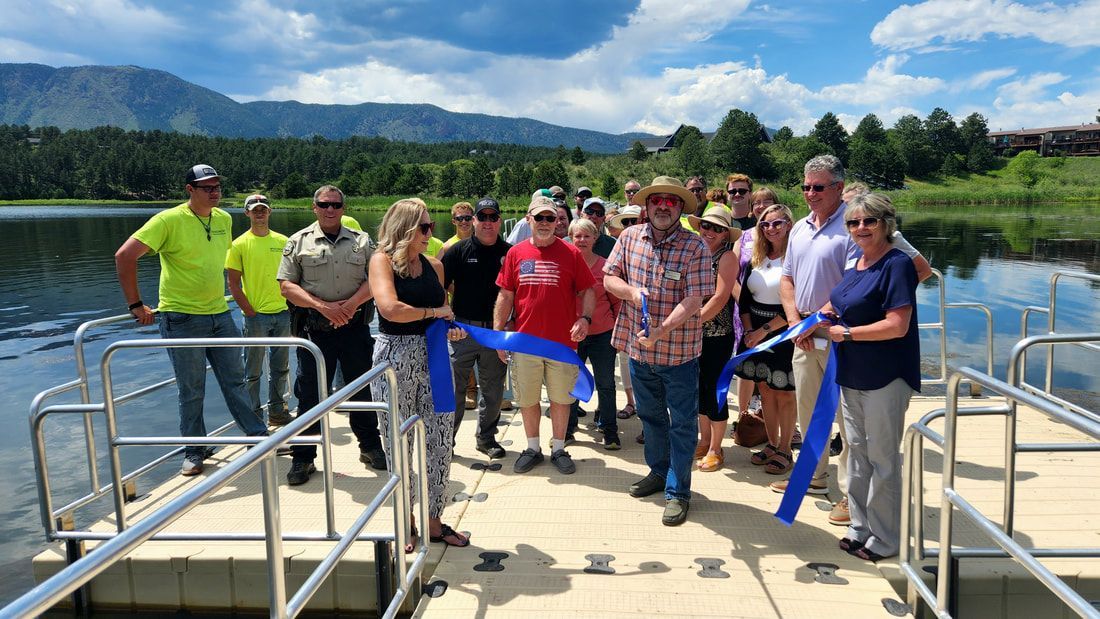 People cutting a blue ribbon on a dock near a lake, with mountains in the background.